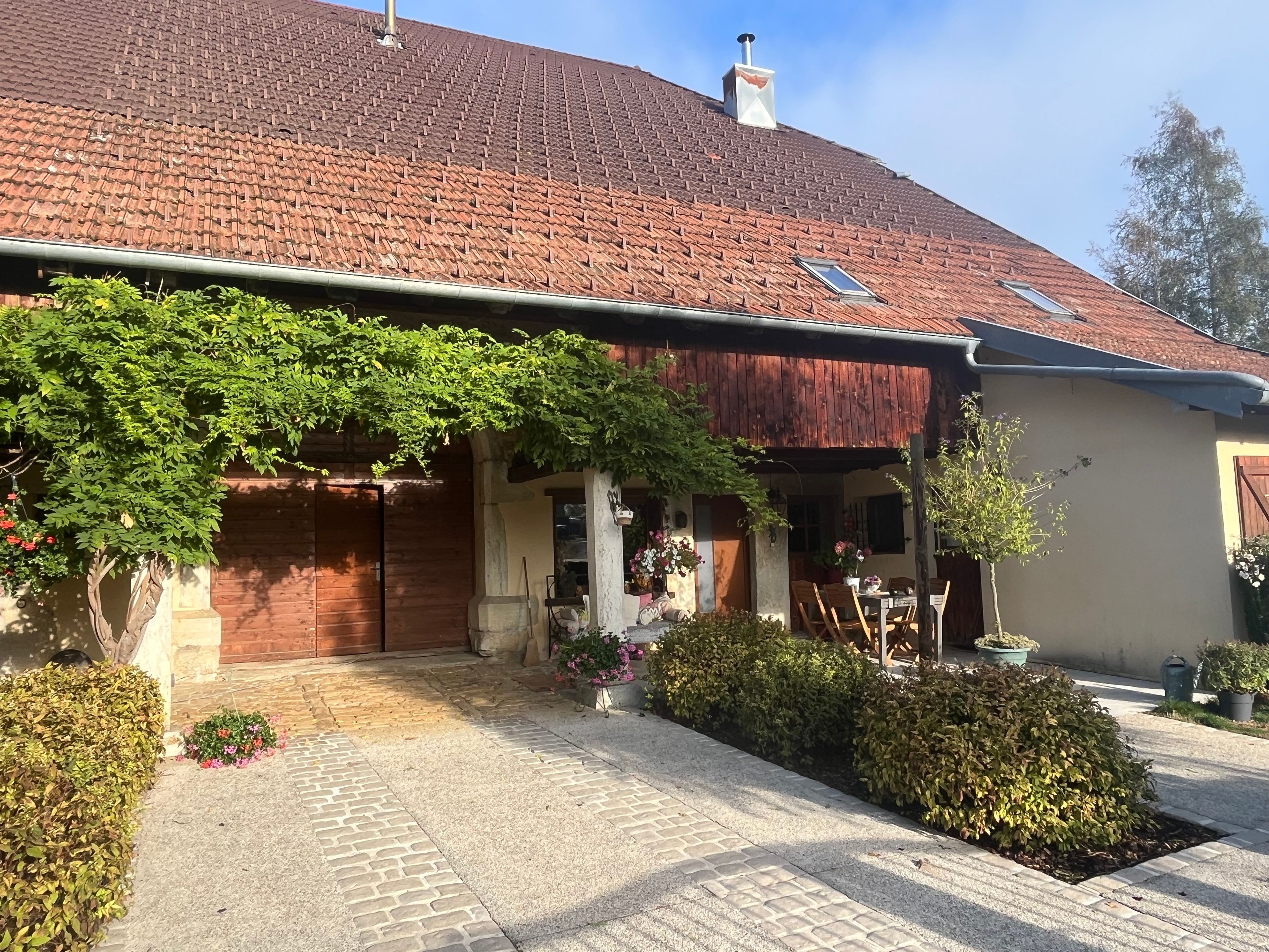 Une maison avec un toit recouvert de tuiles rouges, une véranda ombragée ornée de plantes et une allée pavée.