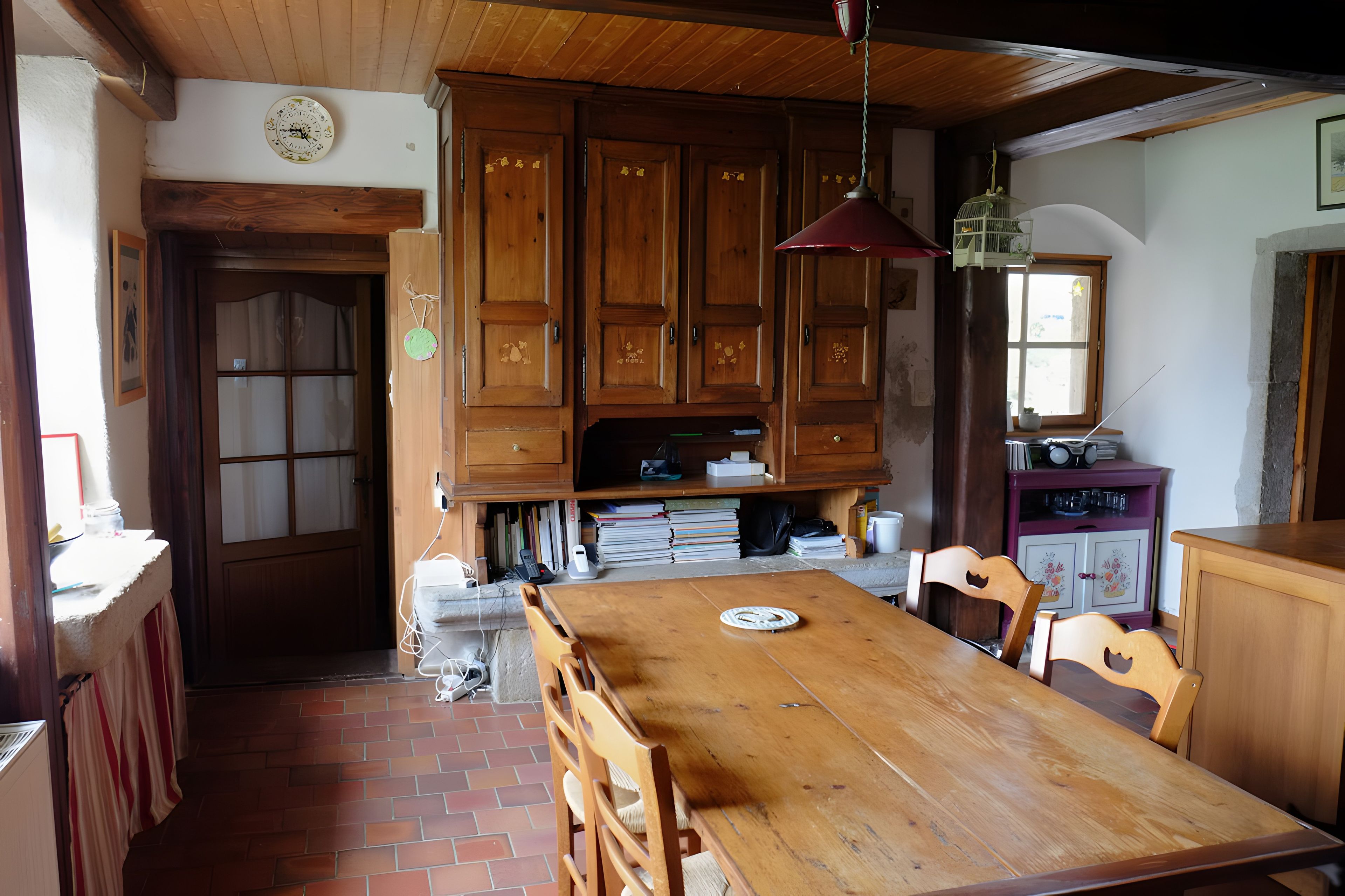 Une table à manger en bois avec des chaises, un coin cuisine avec des placards, une horloge au mur et une fenêtre donnant sur un paysage agréable.