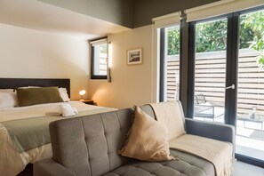 A hotel room with a bed, a sofa, a bedside table with a lamp, a window with blinds, and a view of the outside. - Large Terrace Studio · Selected Design (H02) (Providencia)
