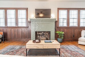 A living room with a brick fireplace, wooden paneled walls, a television mounted above the fireplace, and a potted plant. - The Dapper Duck-Downtown Southern Pines! (Southern Pines)