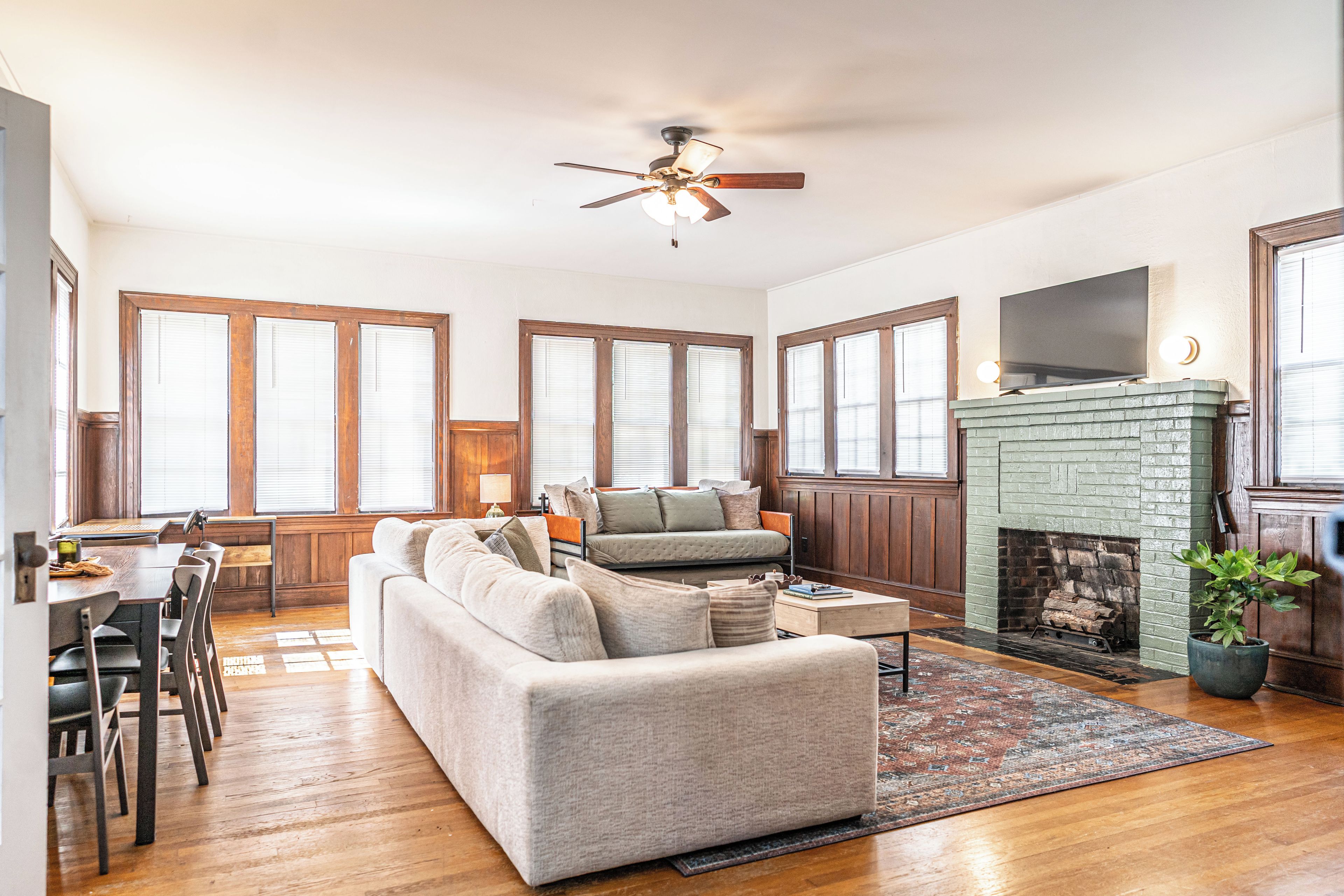 A spacious living room with a fireplace, a dining area with a wooden table and chairs, and large windows with blinds.