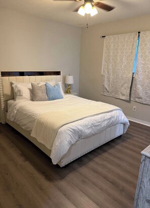 A bedroom with a large bed, a ceiling fan, a nightstand with a lamp, and a window with curtains.