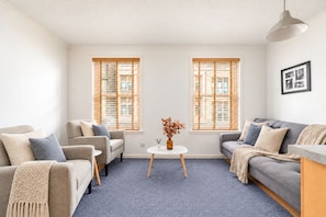 A living room with a grey sofa, a beige armchair, a small white table with a vase, and two windows with wooden blinds. - Charming Edinburgh Flat near Royal Mile (Edinburgh)