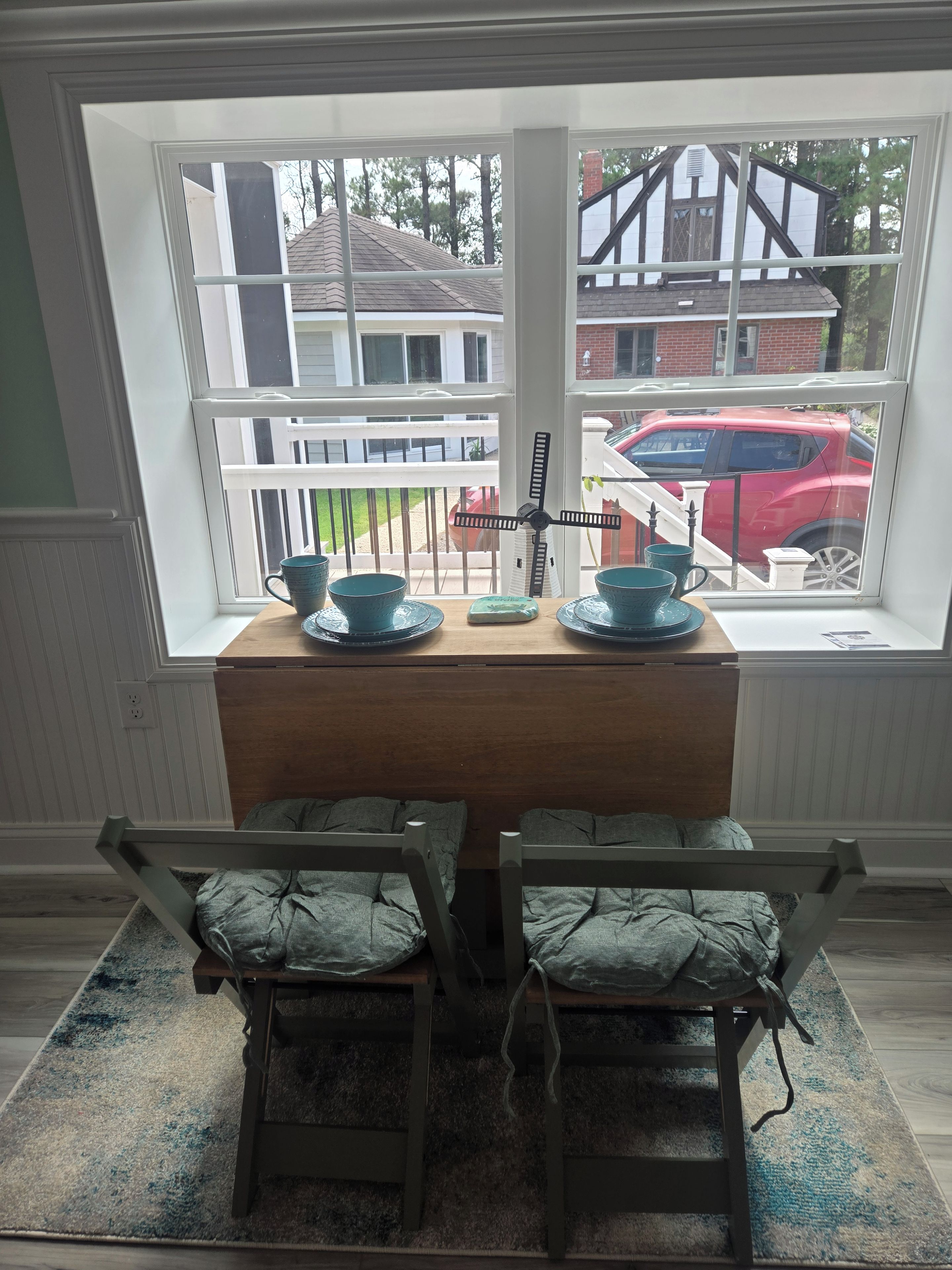 A dining area with a wooden table and chairs, a window with a view of a red car and houses, and a small decorative item on the table.