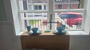 A dining area with a wooden table and chairs, a window with a view of a red car and houses, and a small decorative item on the table.