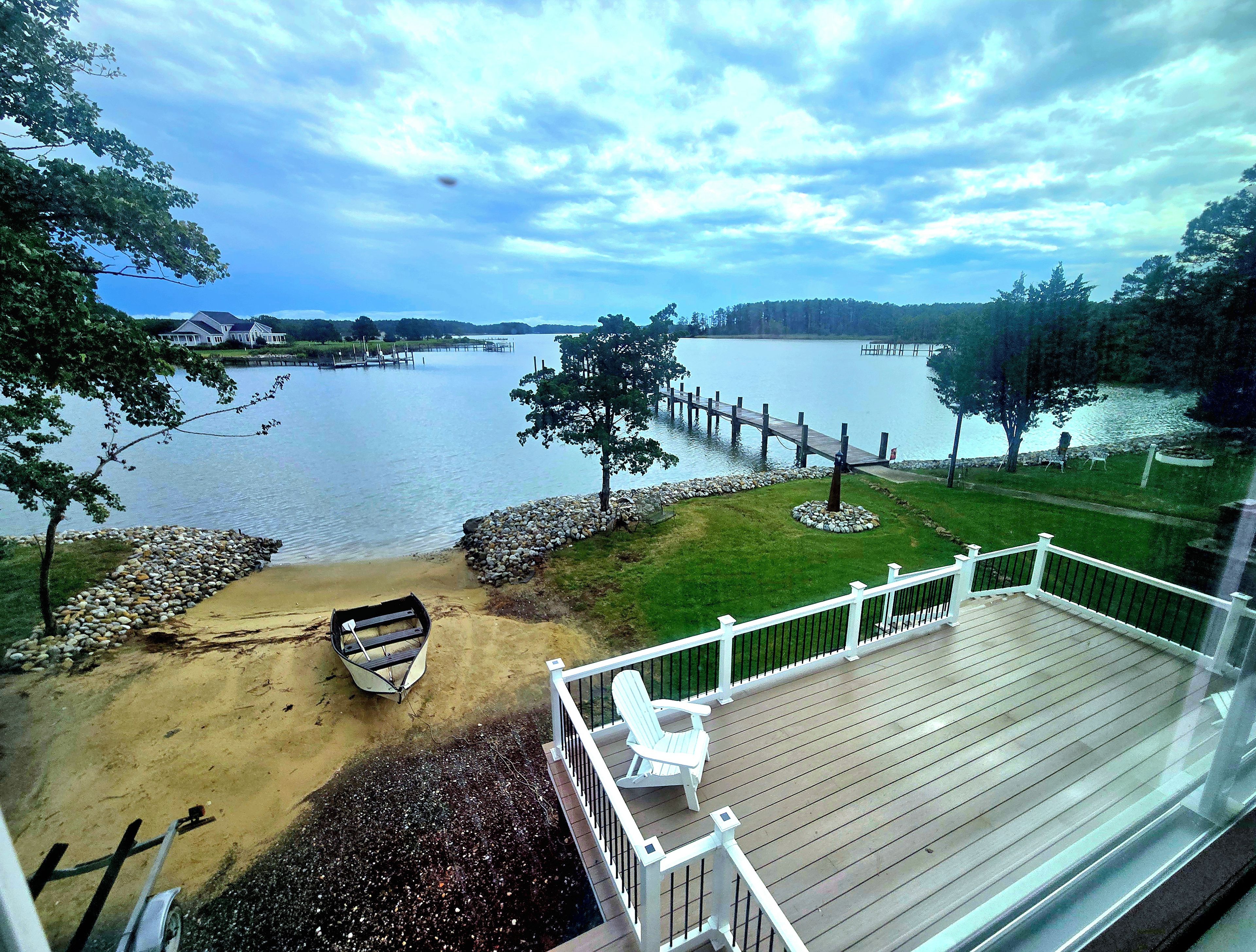 A lakeside view from a deck with a wooden railing, a sandy area with a boat, and a dock extending into the water.