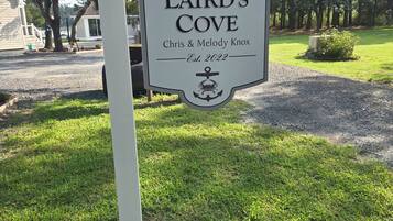 A sign for 'Laird's Cove' established in 2022, honoring Chris & Melody Knox, with a backdrop of trees and a gravel path.