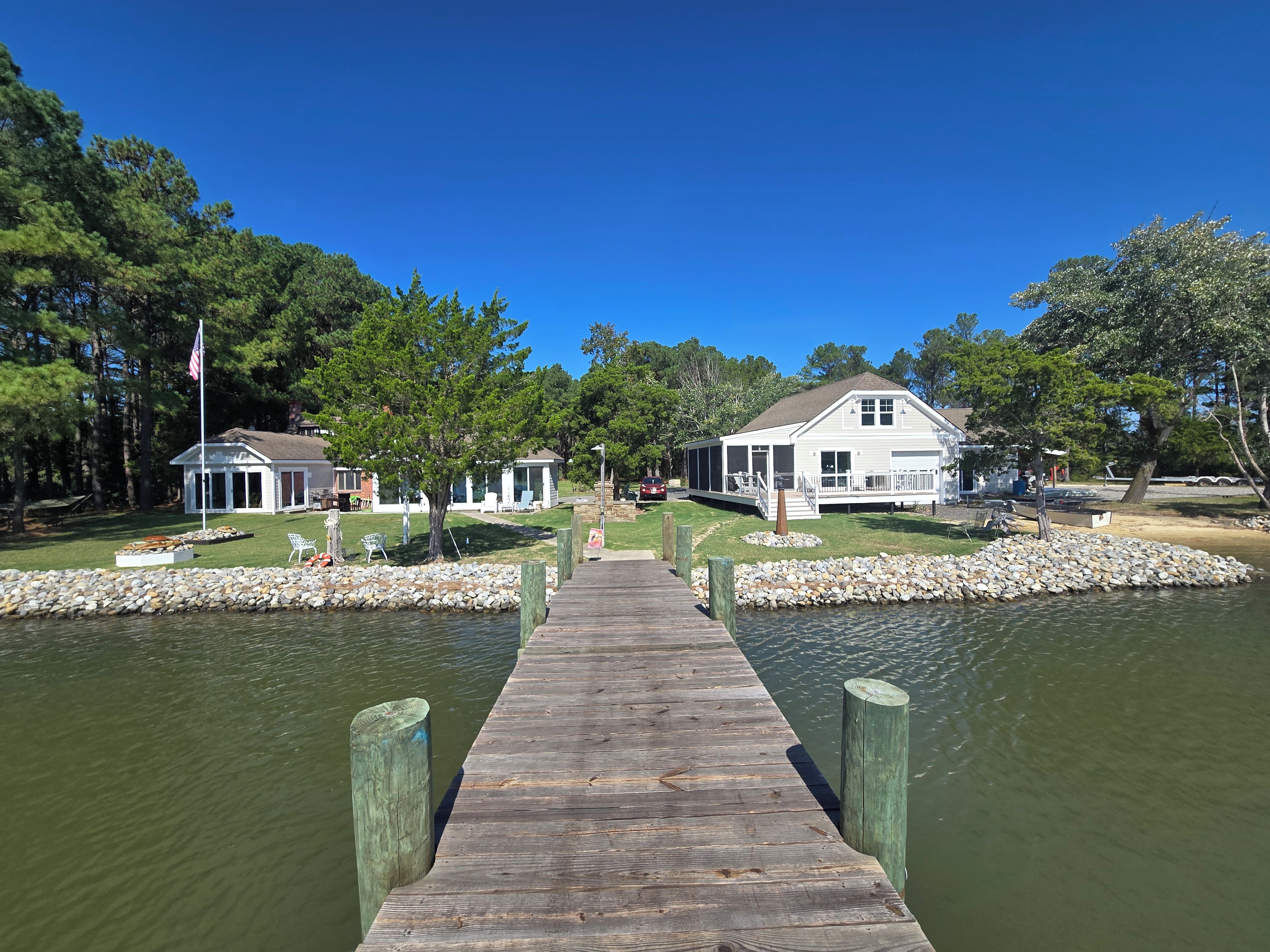 A wooden dock leading to a house with a porch, surrounded by a stone border and trees.
