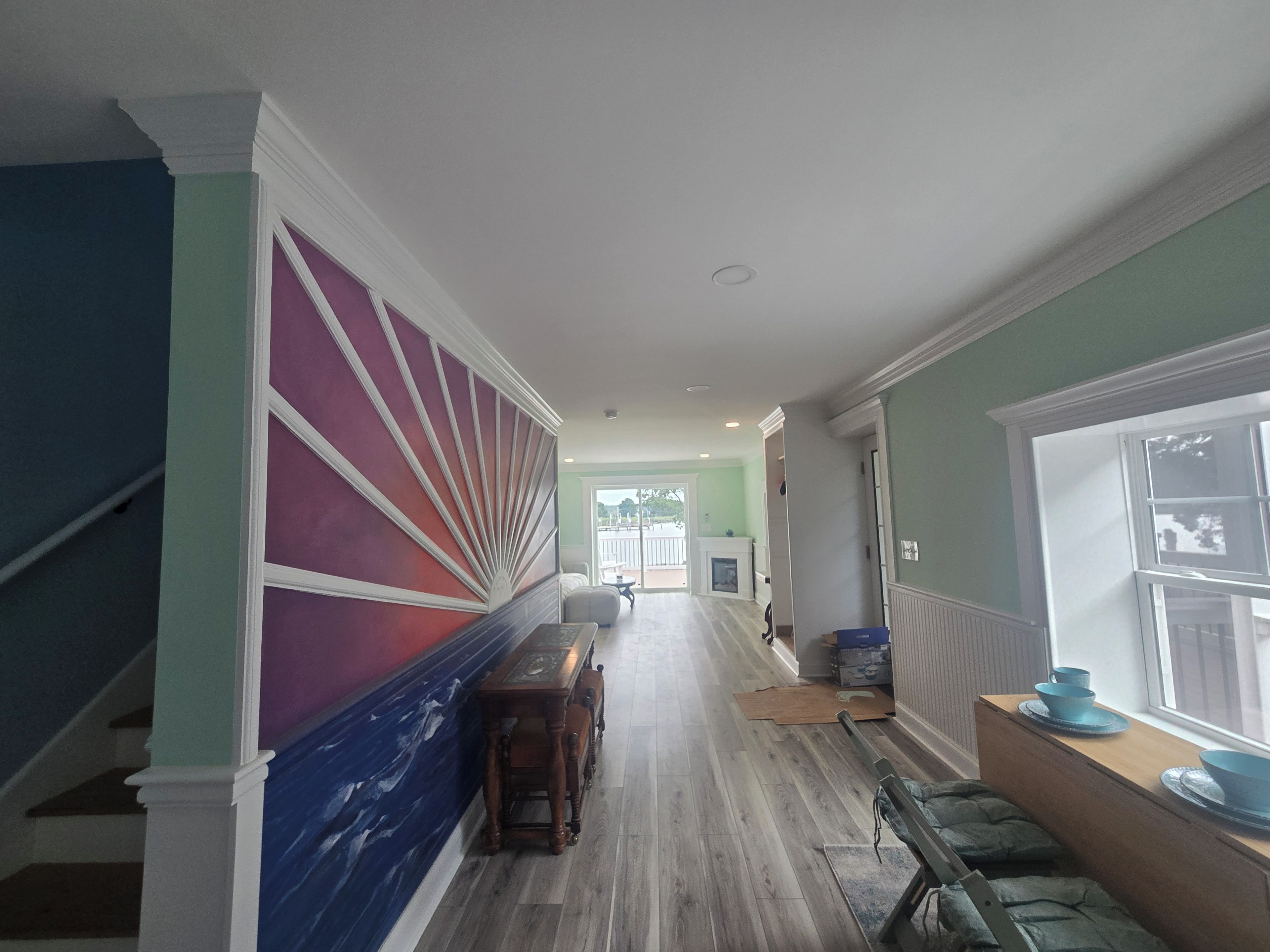 A hallway with a staircase on the left, a dining area with a table and chairs on the right, and a view of the outdoors through a window.