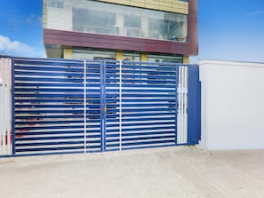Um portão de metal azul com barras horizontais, um edifício com grandes janelas ao fundo e um céu claro.