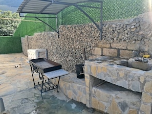 Un área de barbacoa al aire libre con pared de piedra, una parrilla y una mesa de metal.