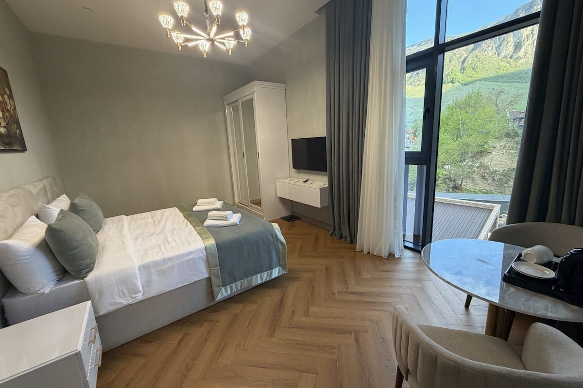 A hotel room with a bed, a desk, a chair, a TV, and a large window with a view of the outdoors.