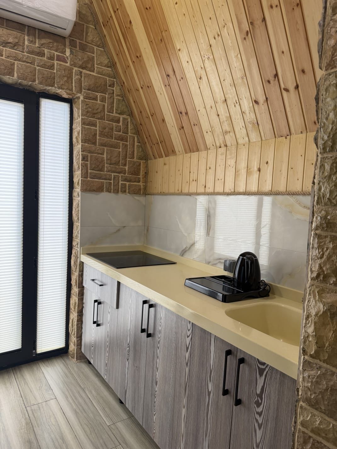 A kitchen with a stone wall, wooden ceiling, and a window with blinds.