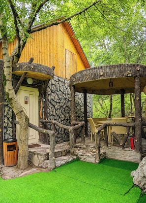 A rustic wooden building with a stone wall, a wooden deck, and a green artificial grass area.