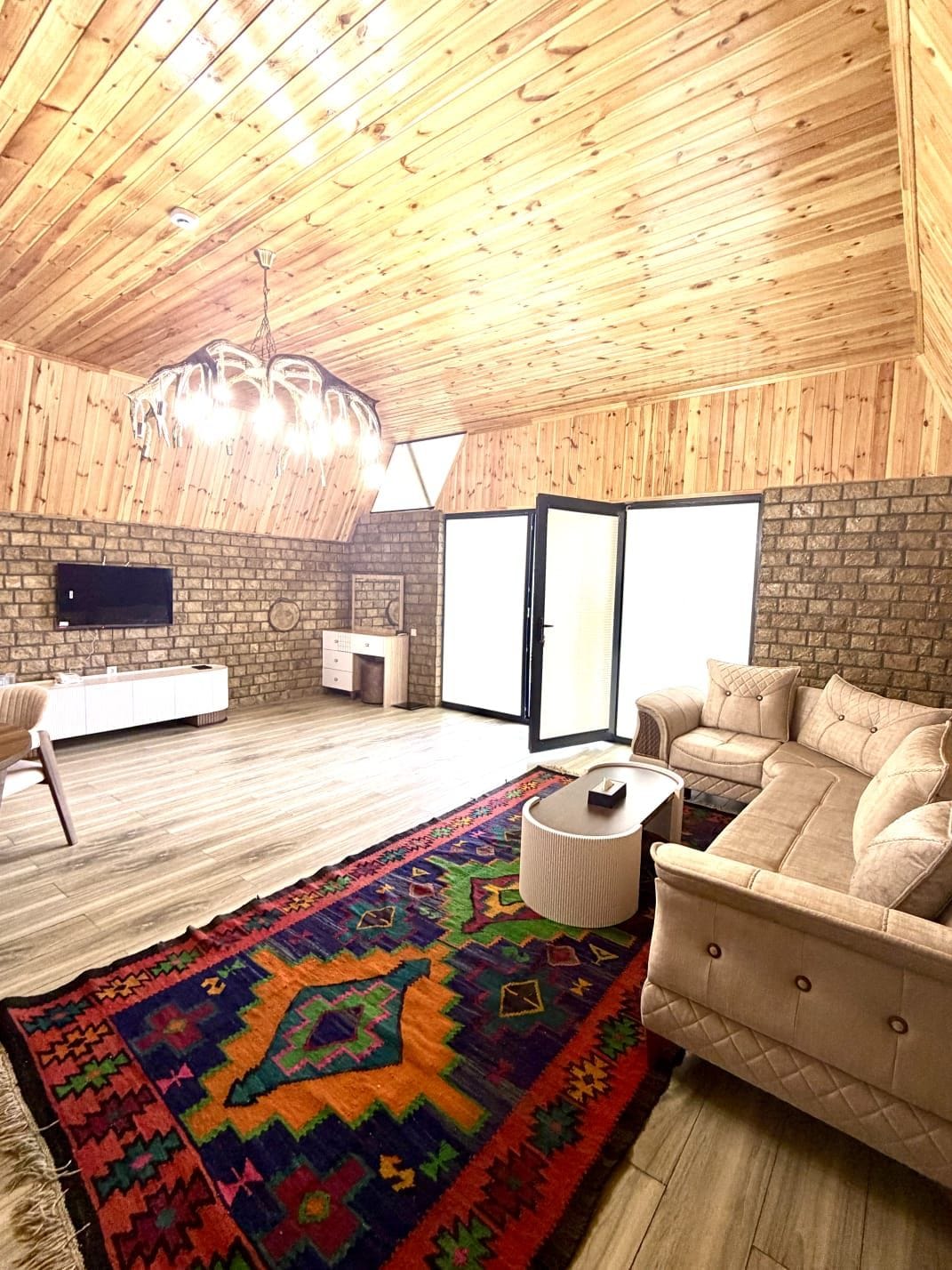 Een ruime kamer met een hoog houten plafond, een grote bank, een kamerbreed tapijt en een televisie bevestigd aan een metselwerk muur.