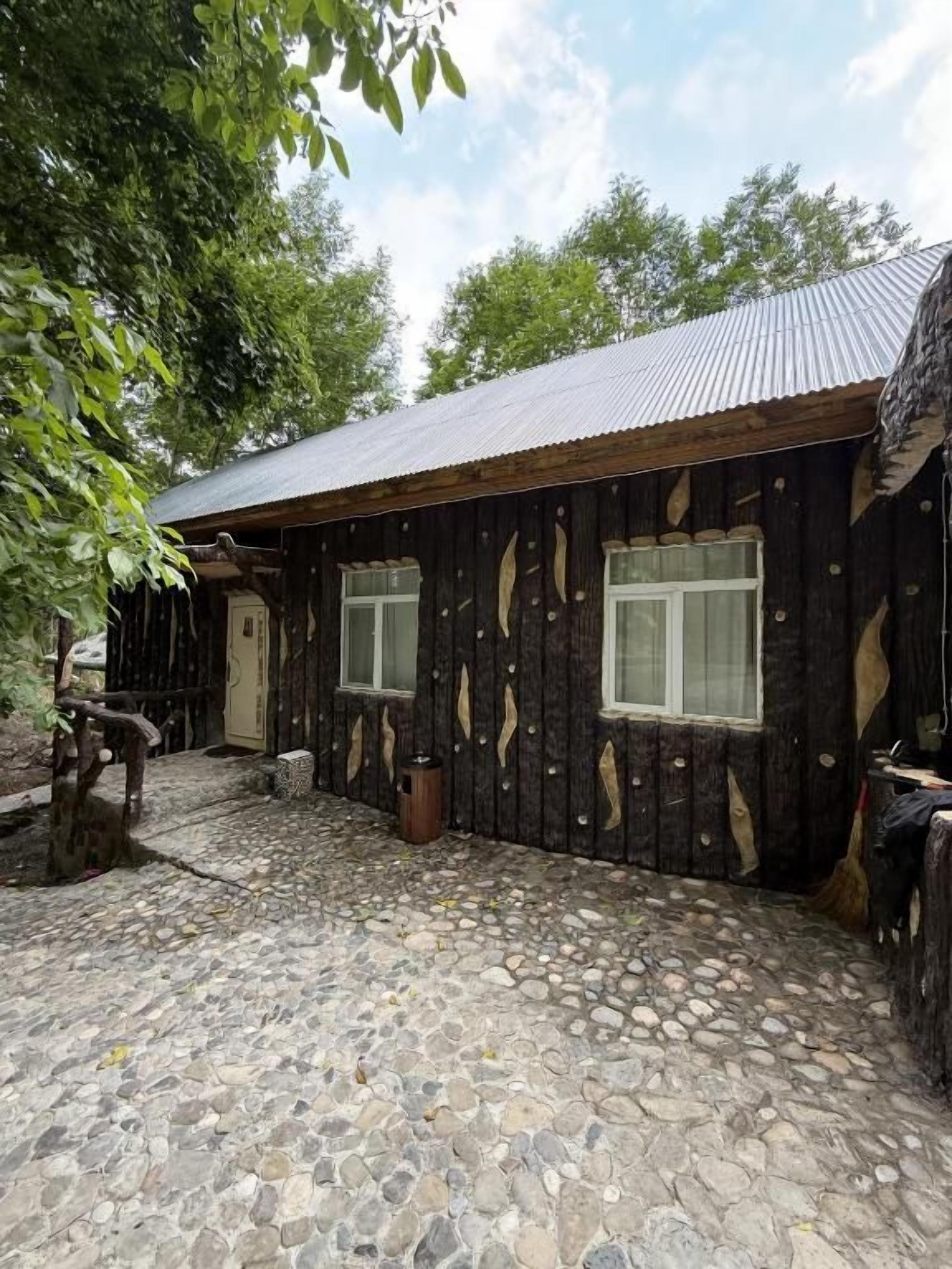 A rustic wooden building with a stone-paved courtyard, a metal roof, and a wooden door.
