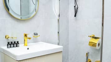 Un baño con tocador blanco, un espejo redondo, un espejo de mano, un secador de pelo y una ducha con manguera dorada.