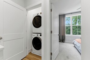 Ein Waschraum mit einer Waschmaschine und einem Trockner, einem Holzboden und einer Tür zu einem Schlafzimmer mit Fenster und Bett.