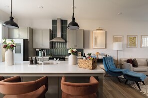 Een moderne keuken met een centraal eiland, hanglampen en een zithoek met een blauwe stoel en poef.
