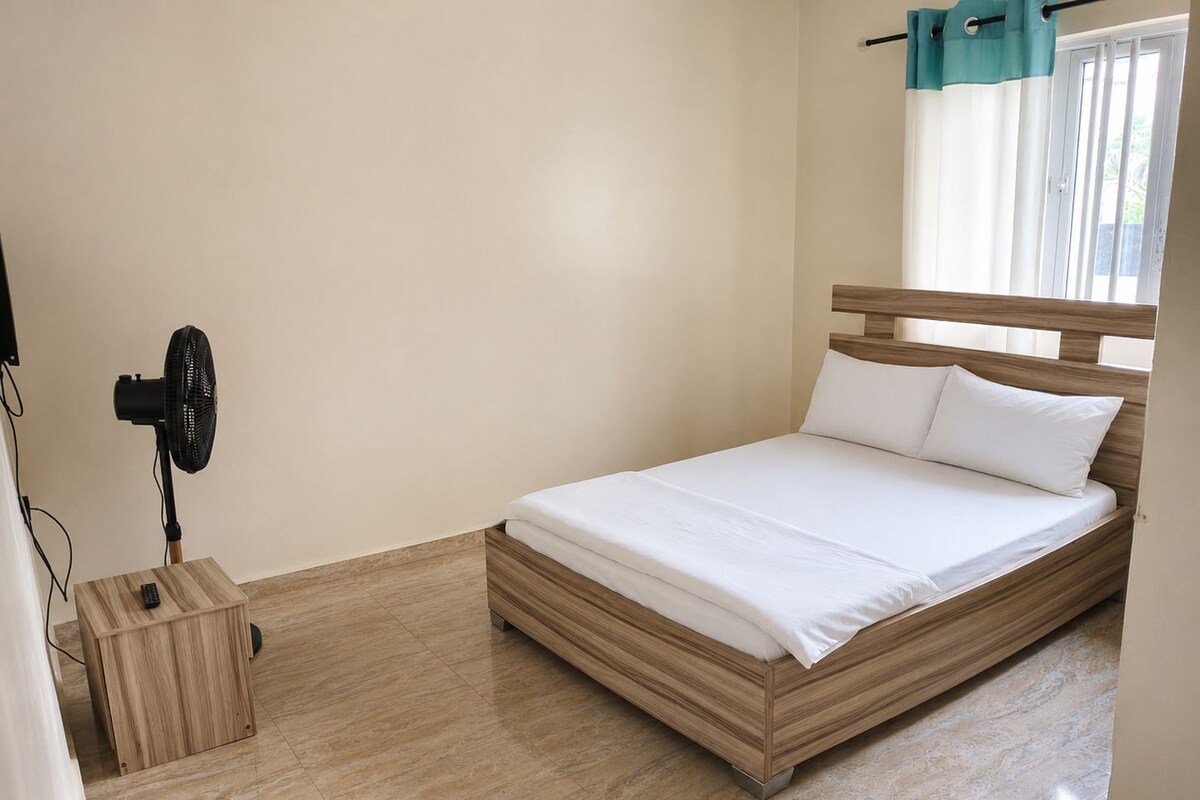 Ein Schlafzimmer mit einem Bett, einem Standventilator, einem Nachttisch aus Holz und einem Fenster mit Vorhängen.