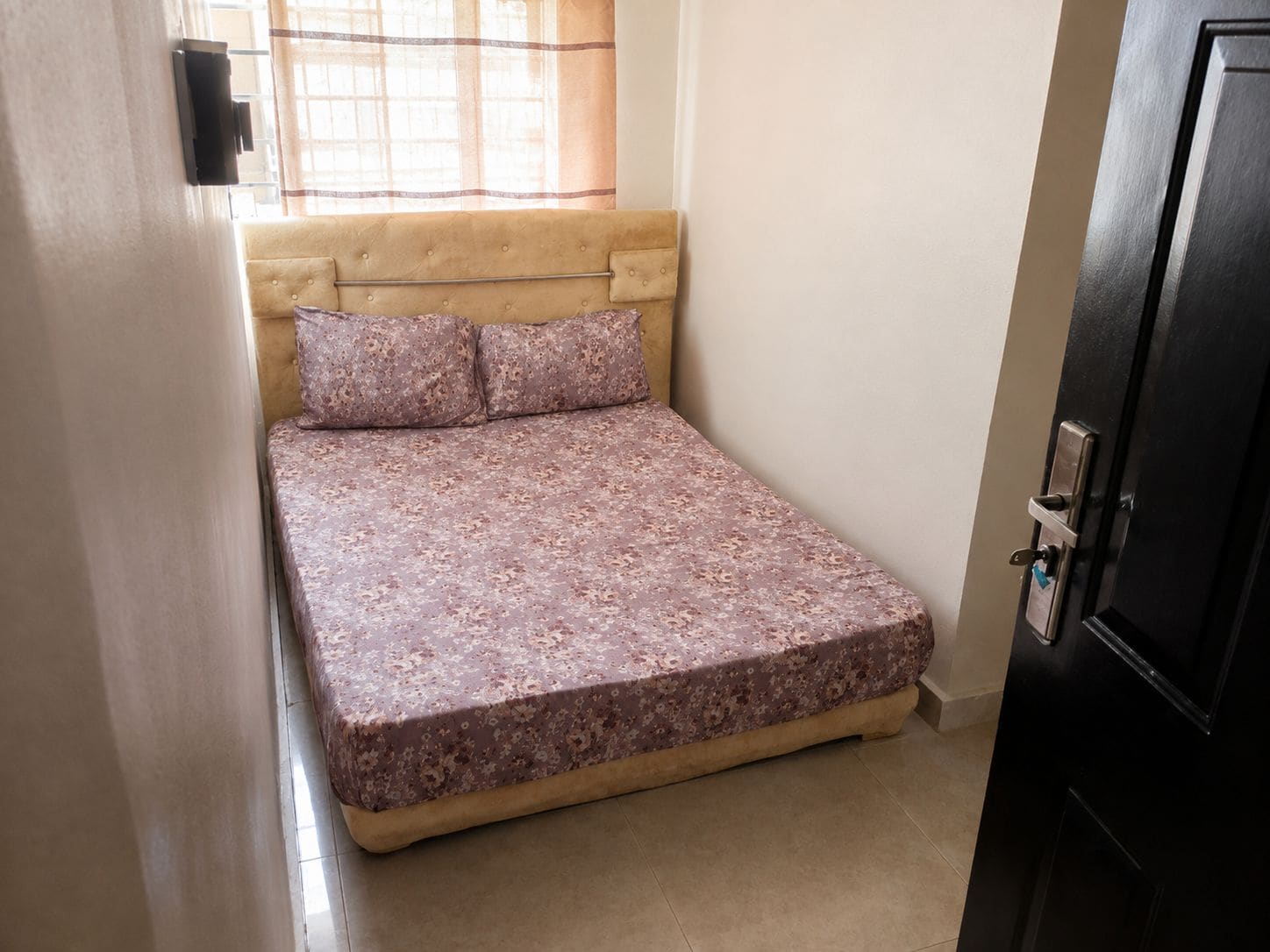 A single bed with a headboard, a window with curtains, and a door with a lock.