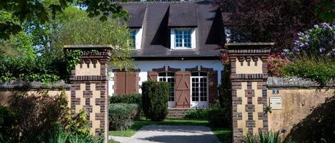 Une maison traditionnelle avec une façade en briques, une porte d’entrée surmontée de volets et une allée pavée menant à l’entrée.
