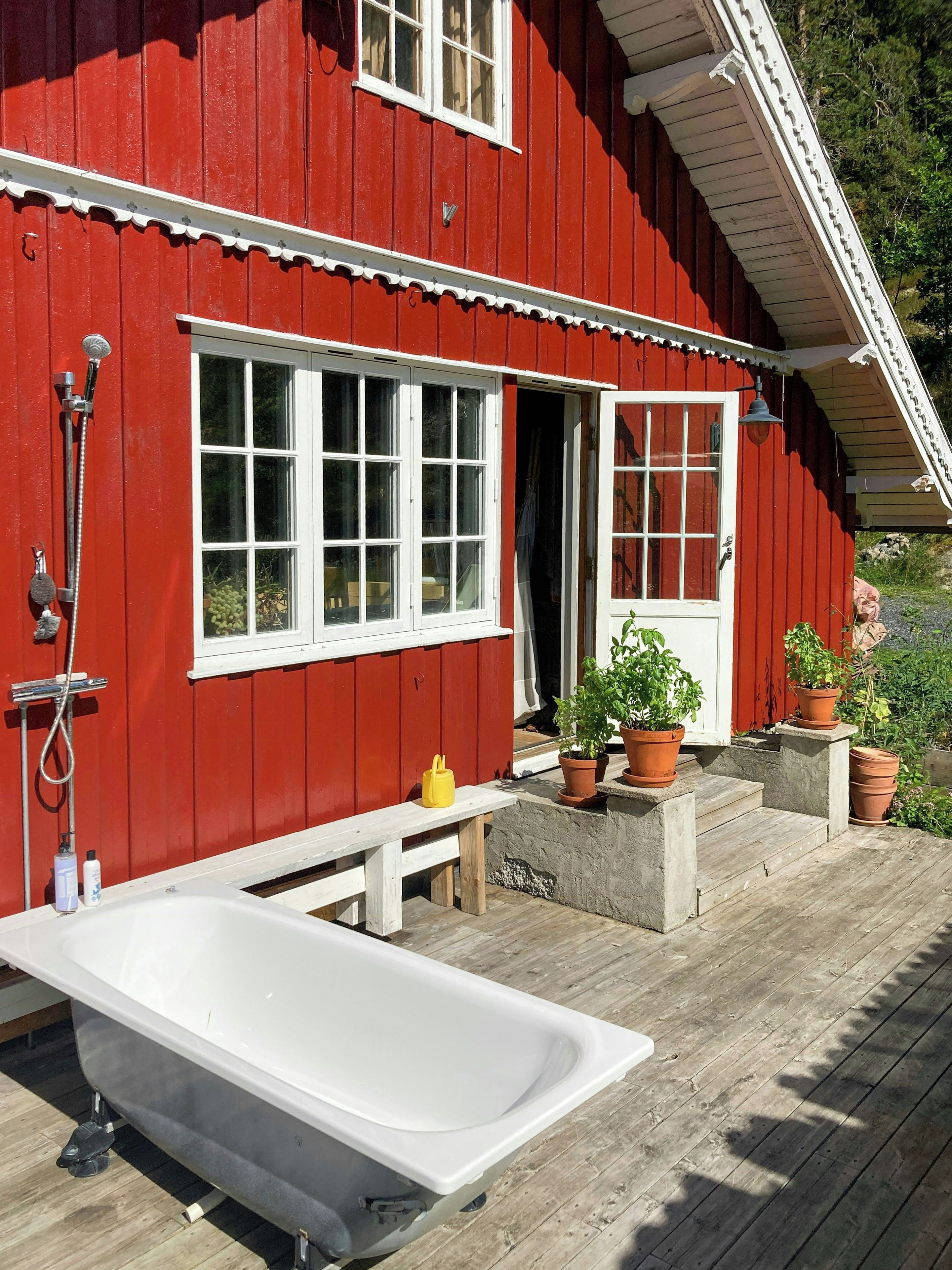 Un bâtiment en bois rouge avec une baignoire blanche, un banc et des plantes en pot sur une terrasse en bois.