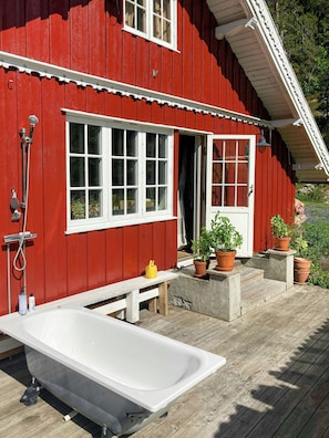 Ein rotes Holzgebäude mit einer weißen Badewanne, einer Bank und Kübelpflanzen auf einer Holzterrasse.