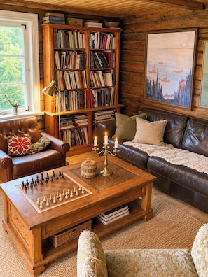 Ein gemütliches Wohnzimmer mit einem Ledersofa, einem Couchtisch mit Schachfiguren, einem Bücherregal und einem Bild an der Wand.