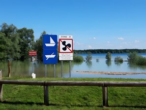 A signpost with directional arrows and a prohibition sign, a wooden fence, and a calm lake with trees in the background.