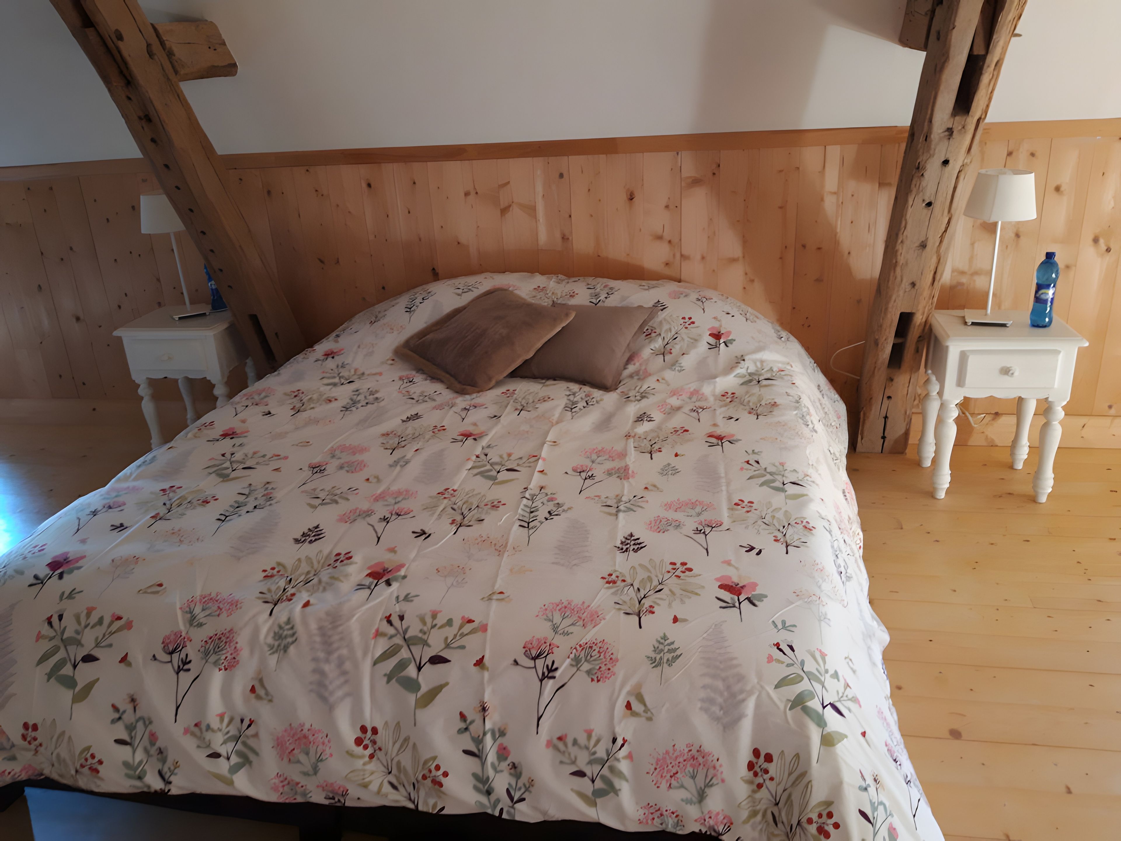 Un dormitorio con colcha estampada de flores, dos almohadas, una mesita de noche con lámpara y techo de madera.