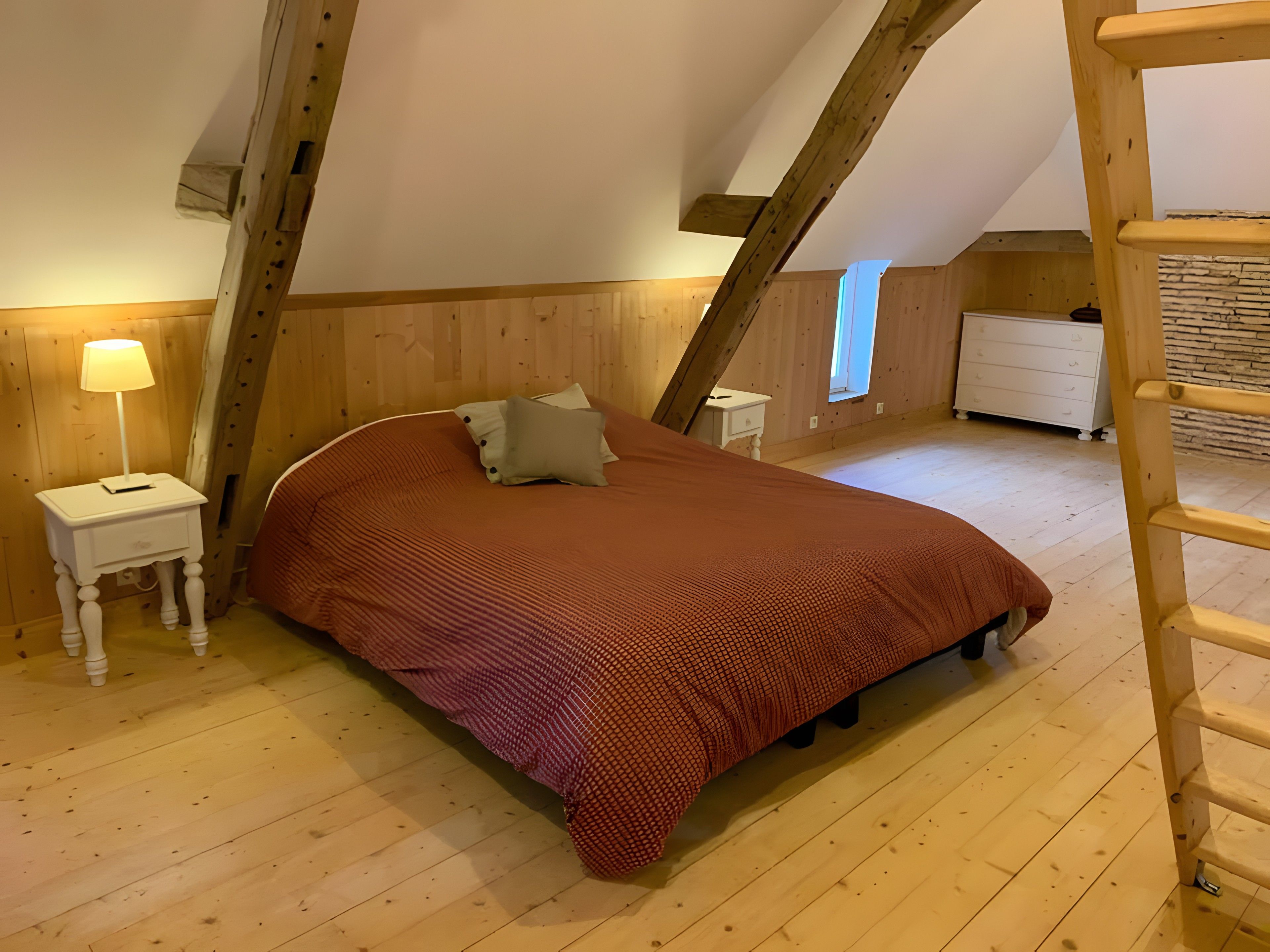 A bedroom with a sloped ceiling, wooden flooring, a bed with a red bedspread, a nightstand with a lamp, a dresser, and a window.