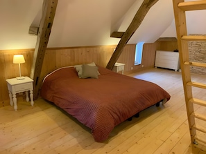 A bedroom with a sloped ceiling, wooden flooring, a bed with a red bedspread, a nightstand with a lamp, a dresser, and a window.