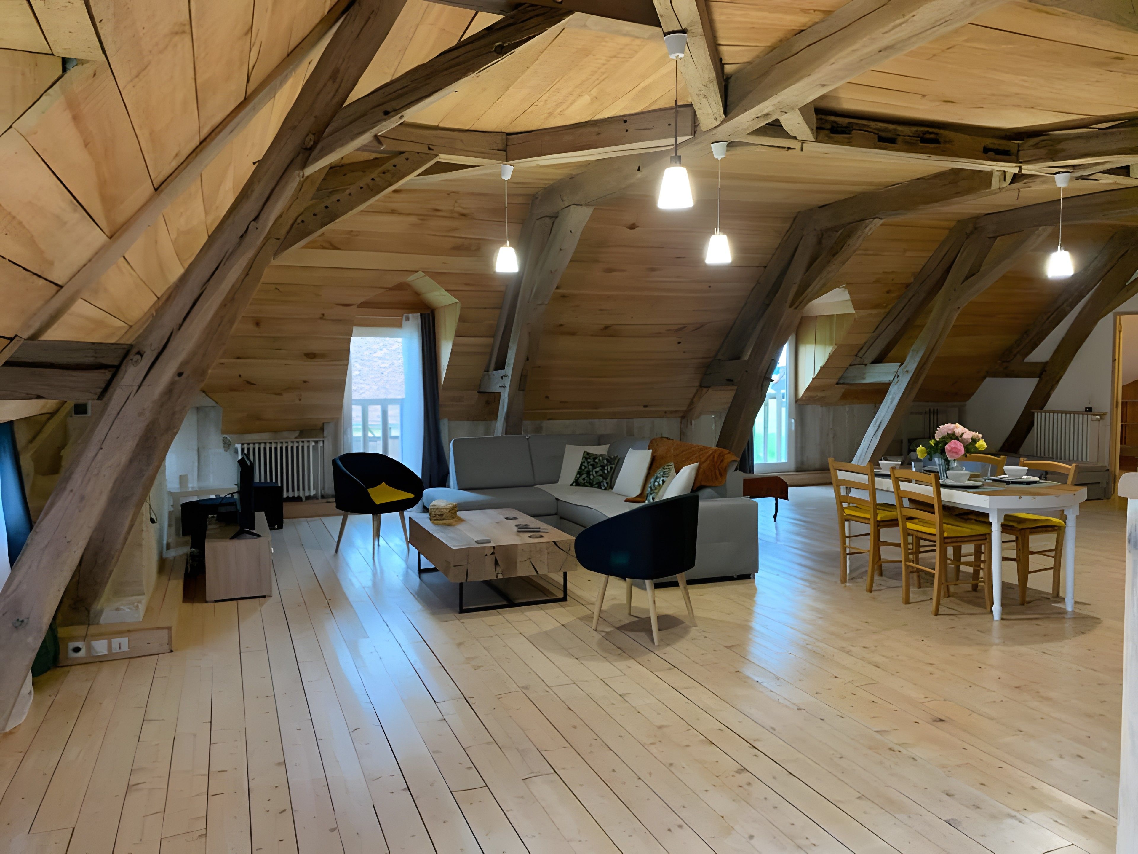 An attic room with exposed wooden beams, a sofa, a dining table with chairs, and pendant lighting.