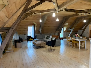 An attic room with exposed wooden beams, a sofa, a dining table with chairs, and pendant lighting.