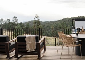 Eine Holzterrasse mit Gartenmöbeln, einem Tisch mit Flasche und Gläsern und Blick auf eine bewaldete Landschaft.