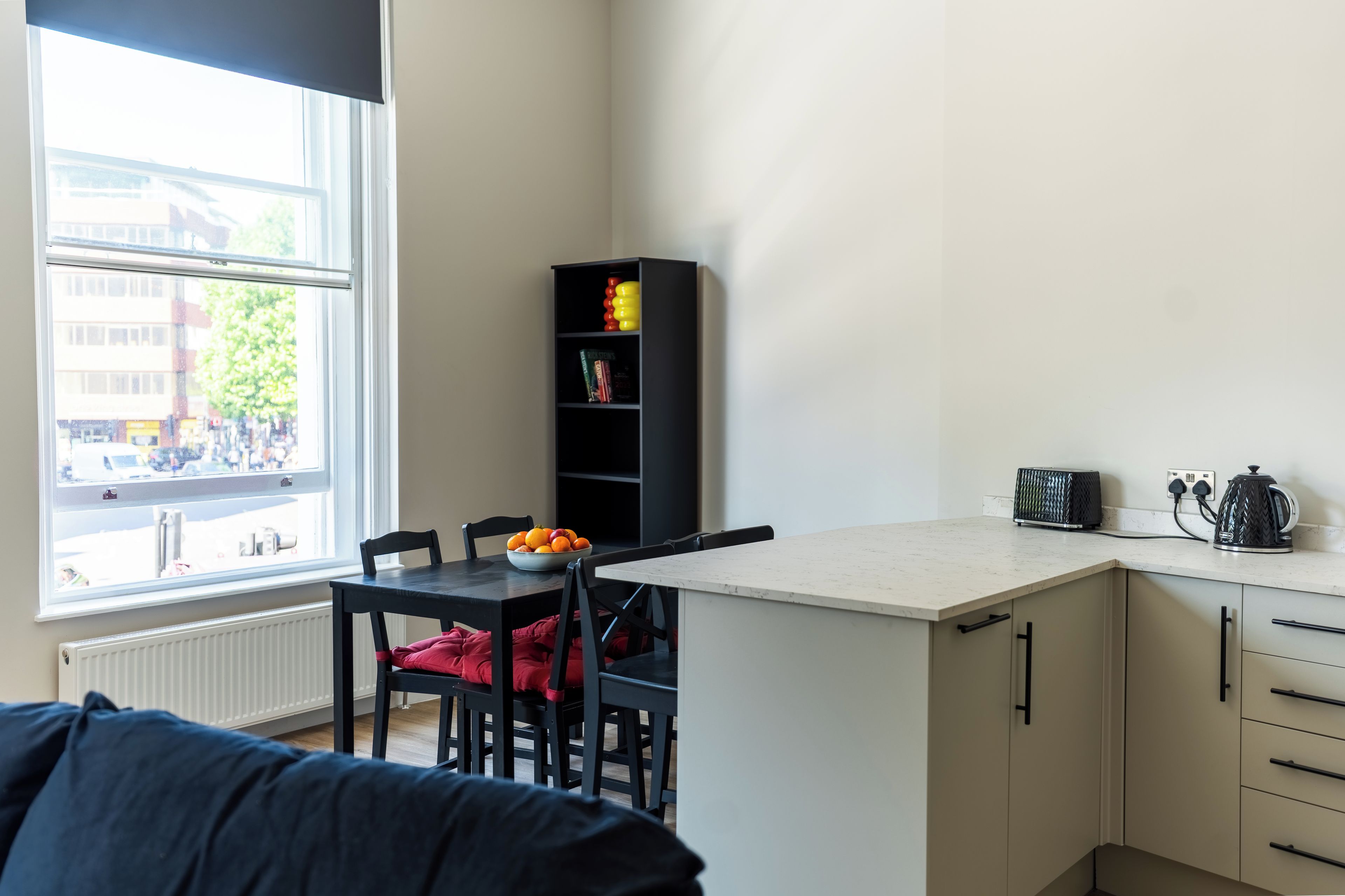 Une petite cuisine équipée avec un coin repas, une fenêtre donnant sur la rue et une bibliothèque noire.