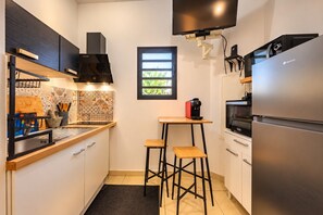 A compact kitchen with a built-in oven, a refrigerator, a coffee machine, and a small dining area with stools. - Hostcare - Studio Piton - Swimming pool & Hiking (Saint-Louis)