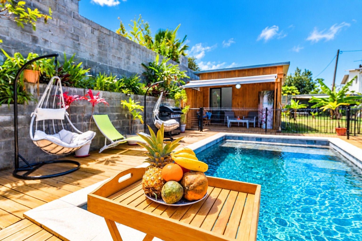 A poolside area with a wooden table, a hanging chair, and a small building with a balcony.