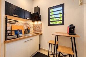 A compact kitchen with a built-in stove, a window with a view of greenery, a coffee machine, and a small stool. - Hostcare - Studio Piton - Swimming pool & Hiking (Saint-Louis)