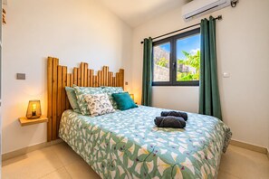 A bedroom with a bed, a bedside lamp, a window with curtains, and an air conditioning unit. - Hostcare - Studio Piton - Swimming pool & Hiking (Saint-Louis)
