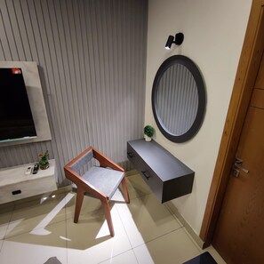 Ein modernes Zimmer mit Fernseher, einem Holzstuhl, einer kleinen Pflanze, einem runden Spiegel und einer grauen Ablagefläche.