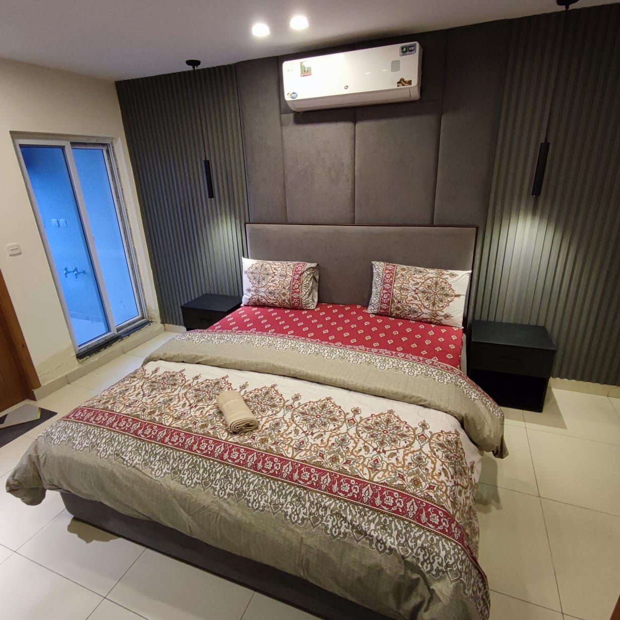 A bedroom with a large bed, a bedside table, a wall-mounted air conditioner, and a window with blinds.