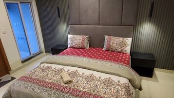 A bedroom with a large bed, a bedside table, a wall-mounted air conditioner, and a window with blinds.