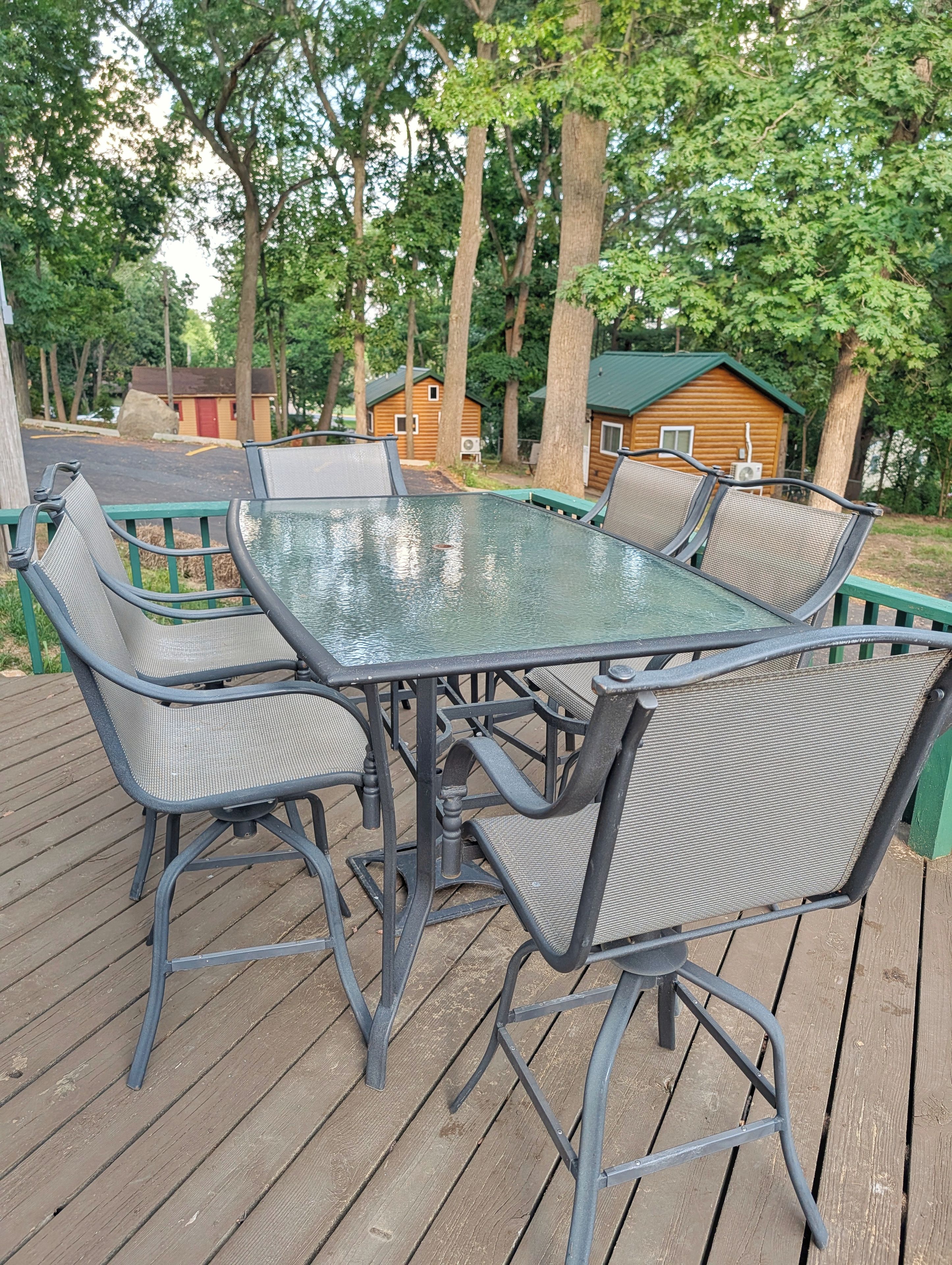 A glass-top table with four chairs on a wooden deck, surrounded by trees and a small cabin in the background.
