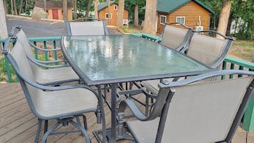 A glass-top table with four chairs on a wooden deck, surrounded by trees and a small cabin in the background.