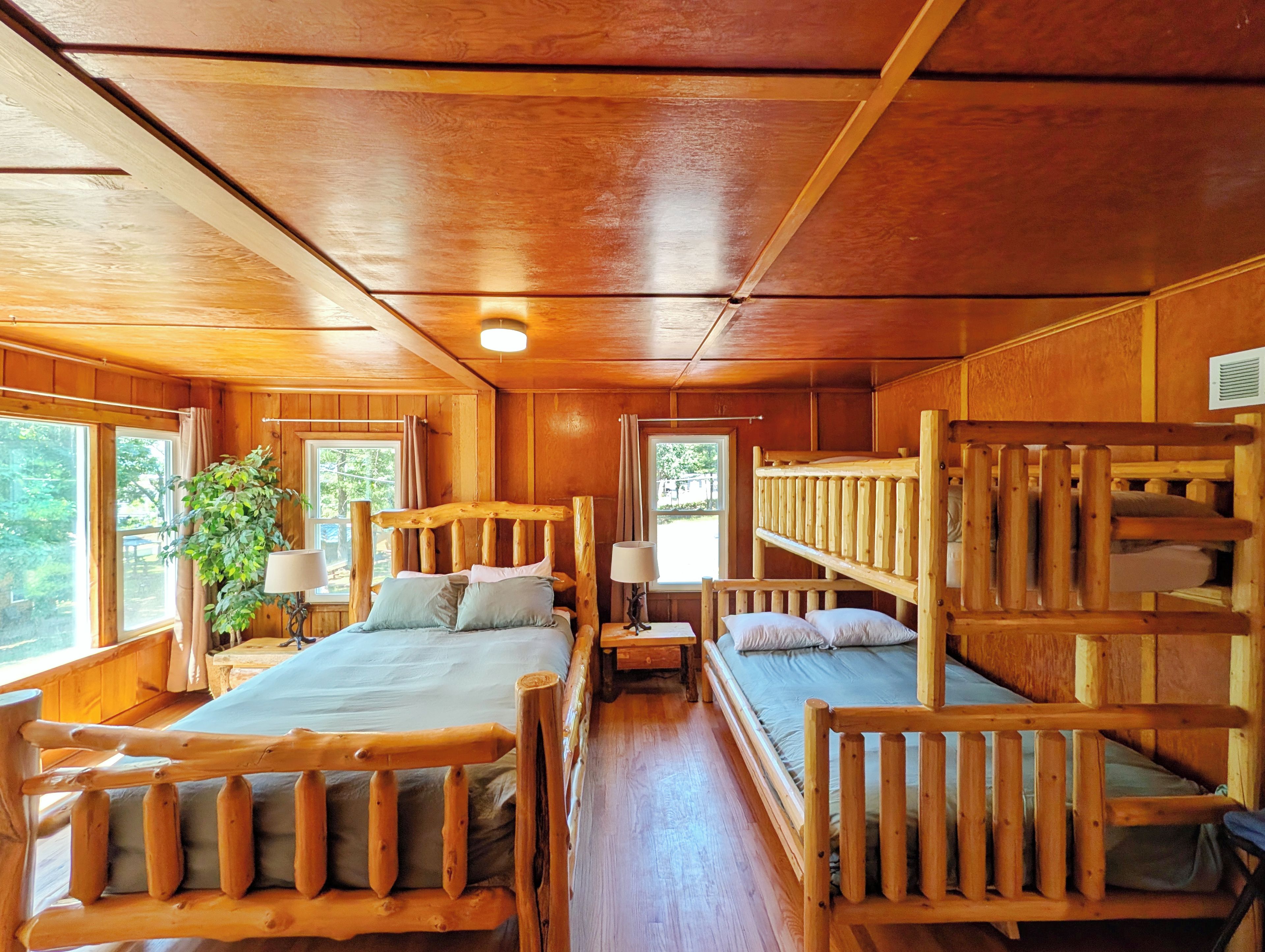 A cabin interior with a wooden bunk bed, a single bed, a small table, and a lamp.