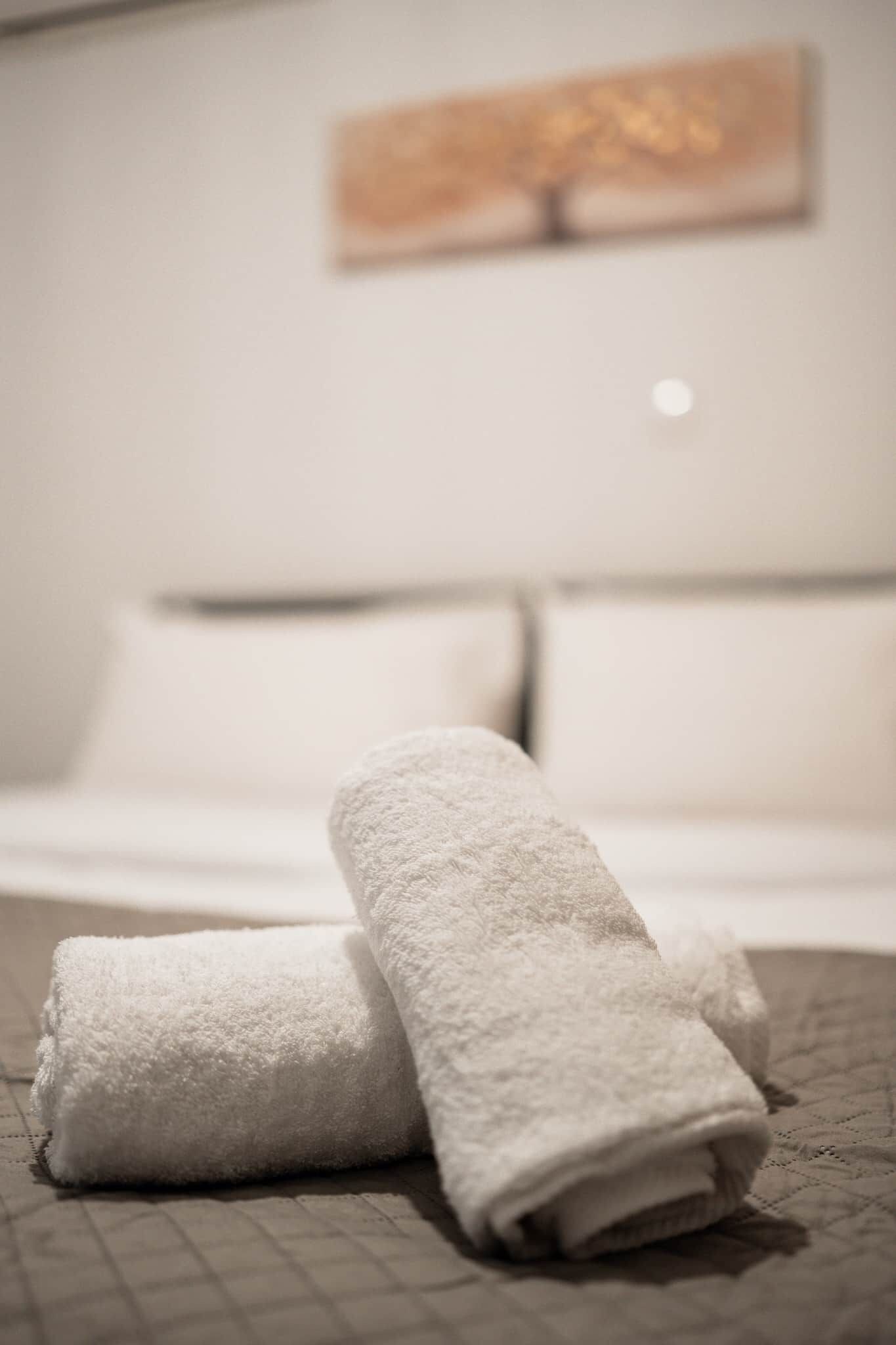 Cama bem arrumada com lençóis brancos e duas toalhas enroladas sobre ela.