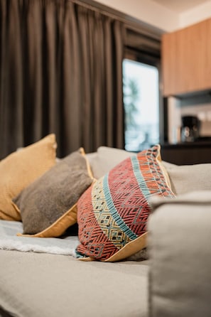 Una cama con una manta y almohadas estampadas, una ventana con cortinas y una cocina al fondo.