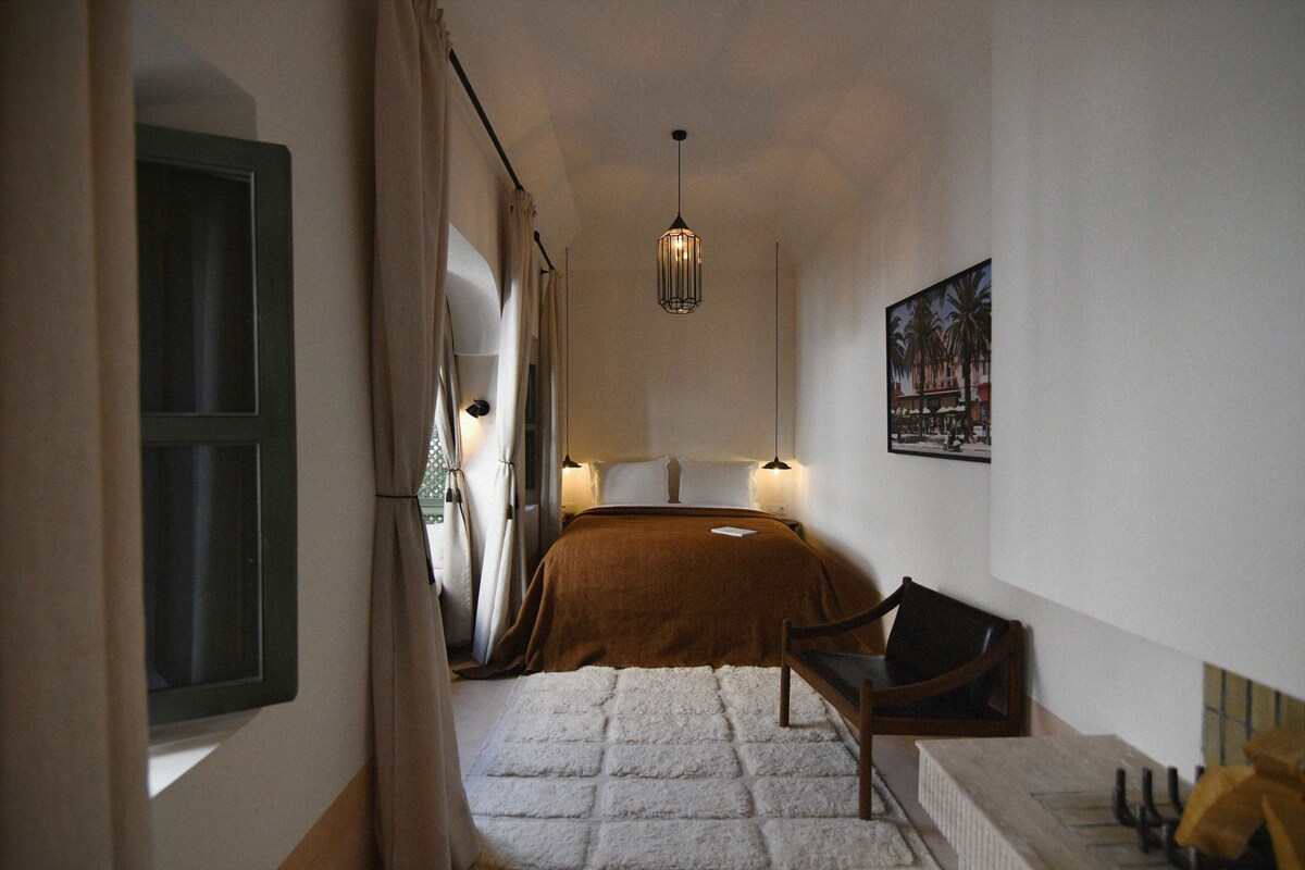 A narrow hallway leading to a bedroom with a bed, a chair, and a painting on the wall.